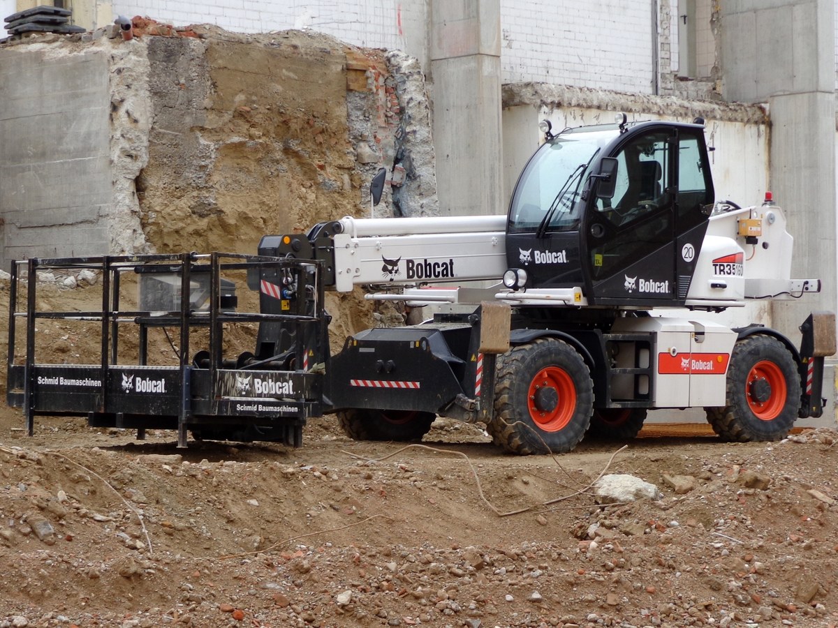 White Bobcat rotating telehandler with outriggers working beside a masonry wall